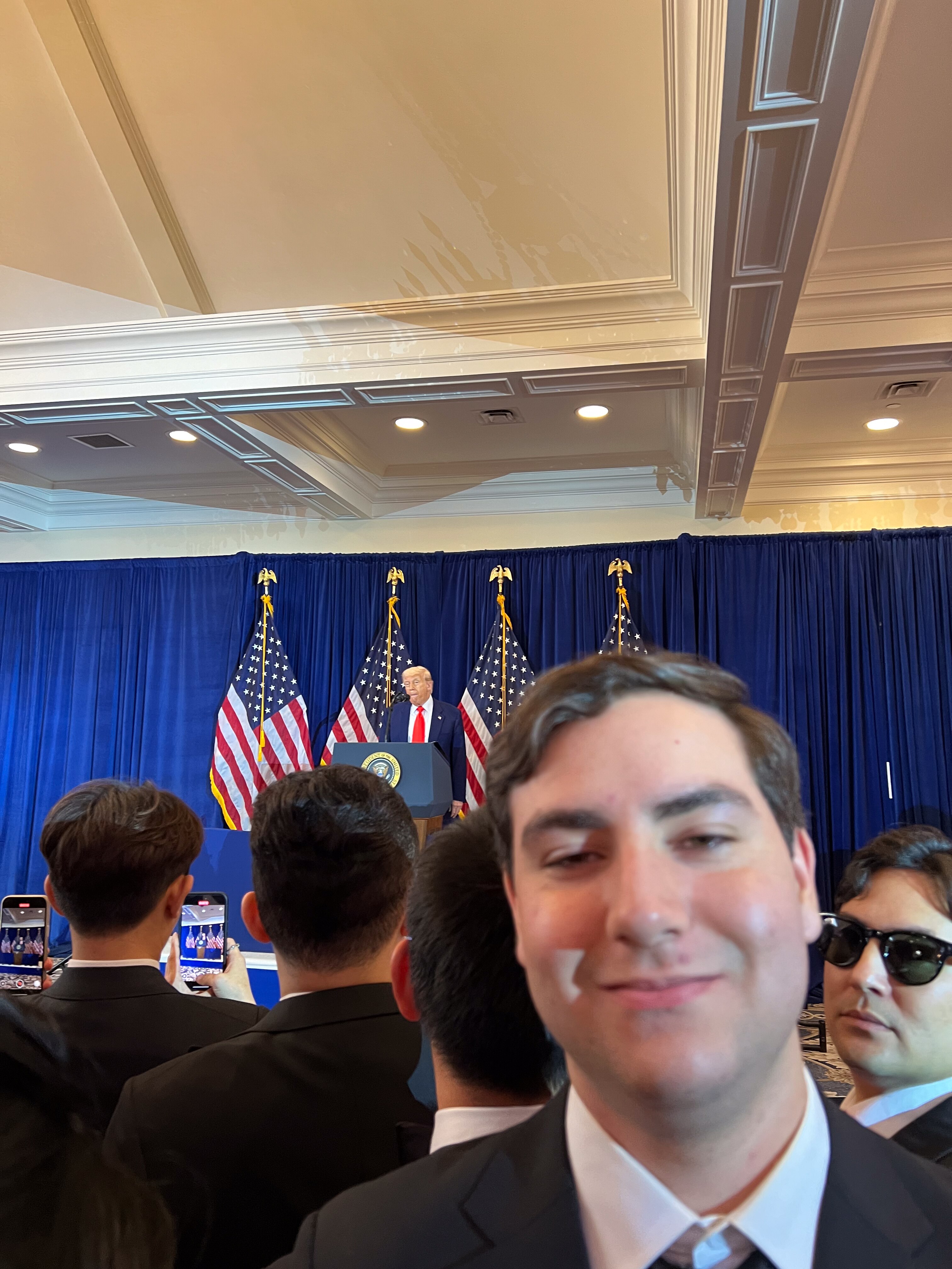 A young man wearing a suit and tie takes a selfie while Donald Trump speaks on stage at a private event.