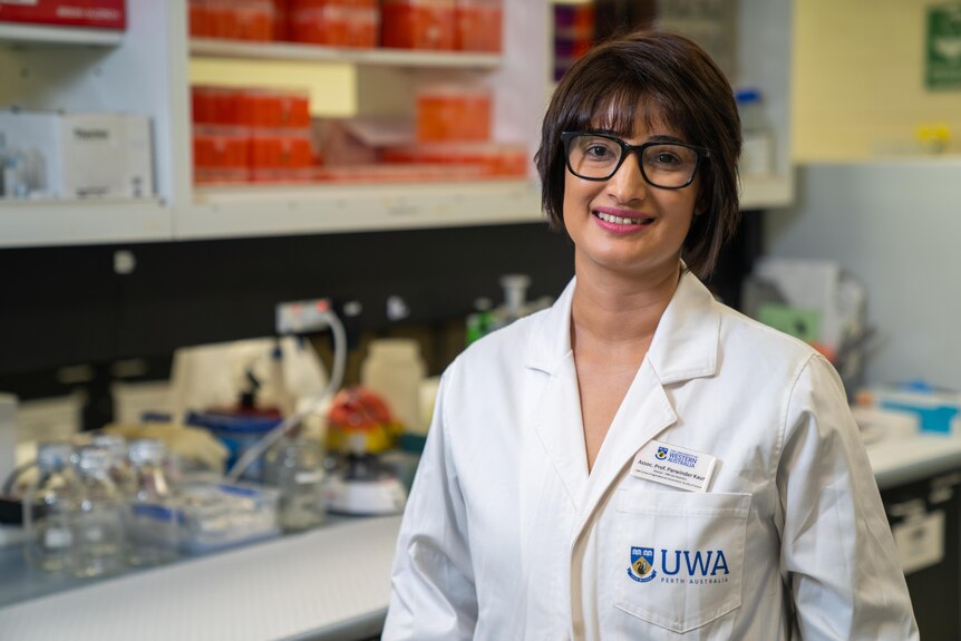 A smiling, dark-haired woman wearing glasses and a white coat stands in a laboratory.