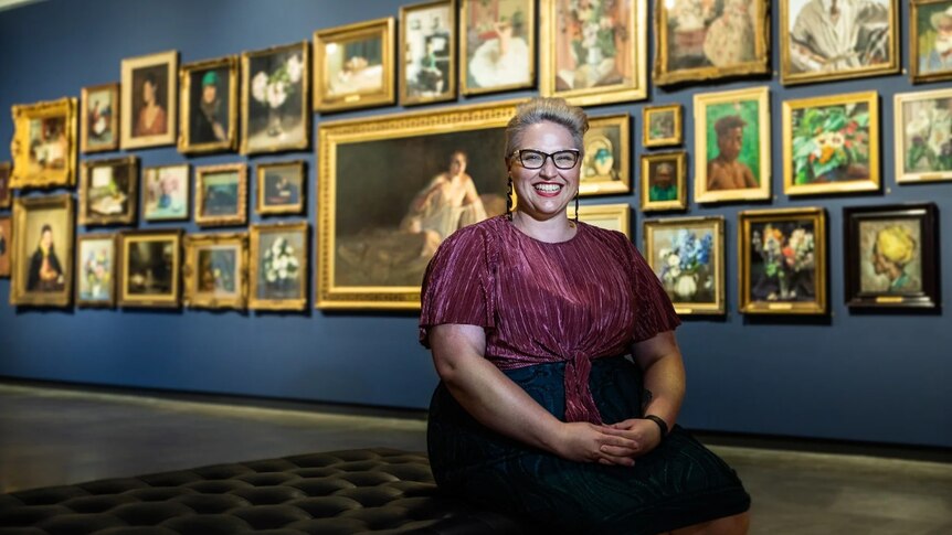 Woman sits smiling on a couch inside an art gallery, with wall of framed artworks behind her.