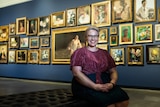 Woman sits smiling on a couch inside an art gallery, with wall of framed artworks behind her.