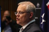 Scott Morrison's lips are pursed  as he speaks. Brendan Murphy is out of focus in the background at his own lectern.