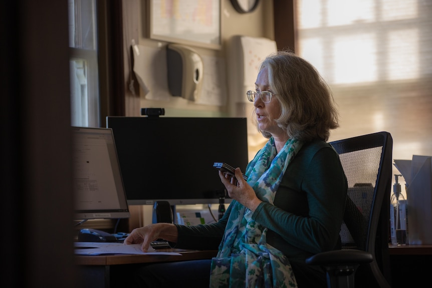 A woman with sliver hair and glasses sits at a desk talking on a smartphone