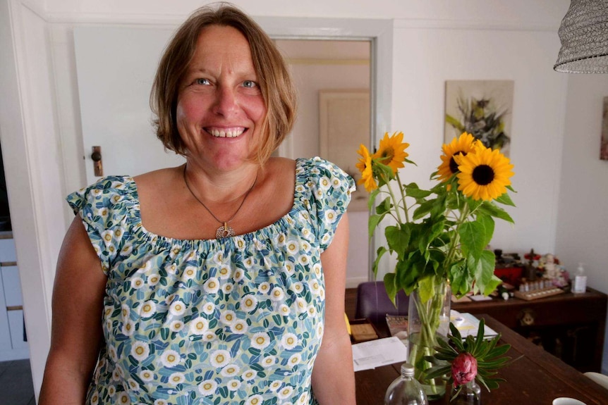 Woman stands near a bunch or bright sunflowers on her kitchen table, smiling in her floral blue blouse.
