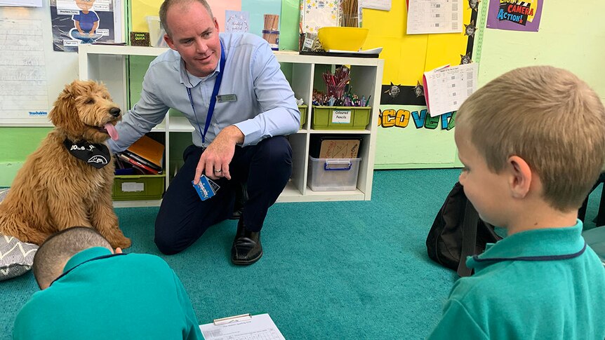 Man kneels on ground with dog next to him looking at primary school aged student