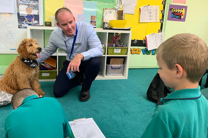 Man kneels on ground with dog next to him looking at primary school aged student