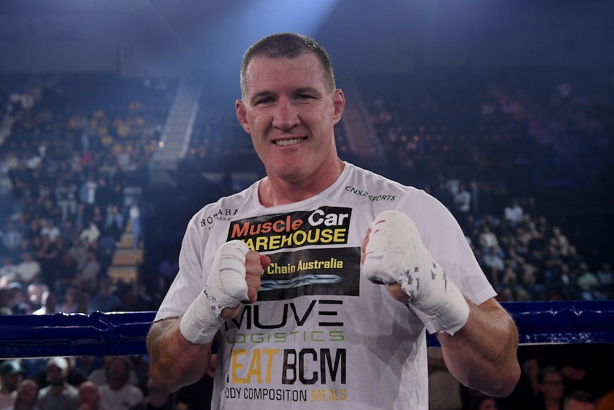 Boxer Paul Gallen smiles at the camera standing in the ring wearing a T-shirt with his hands wrapped. 