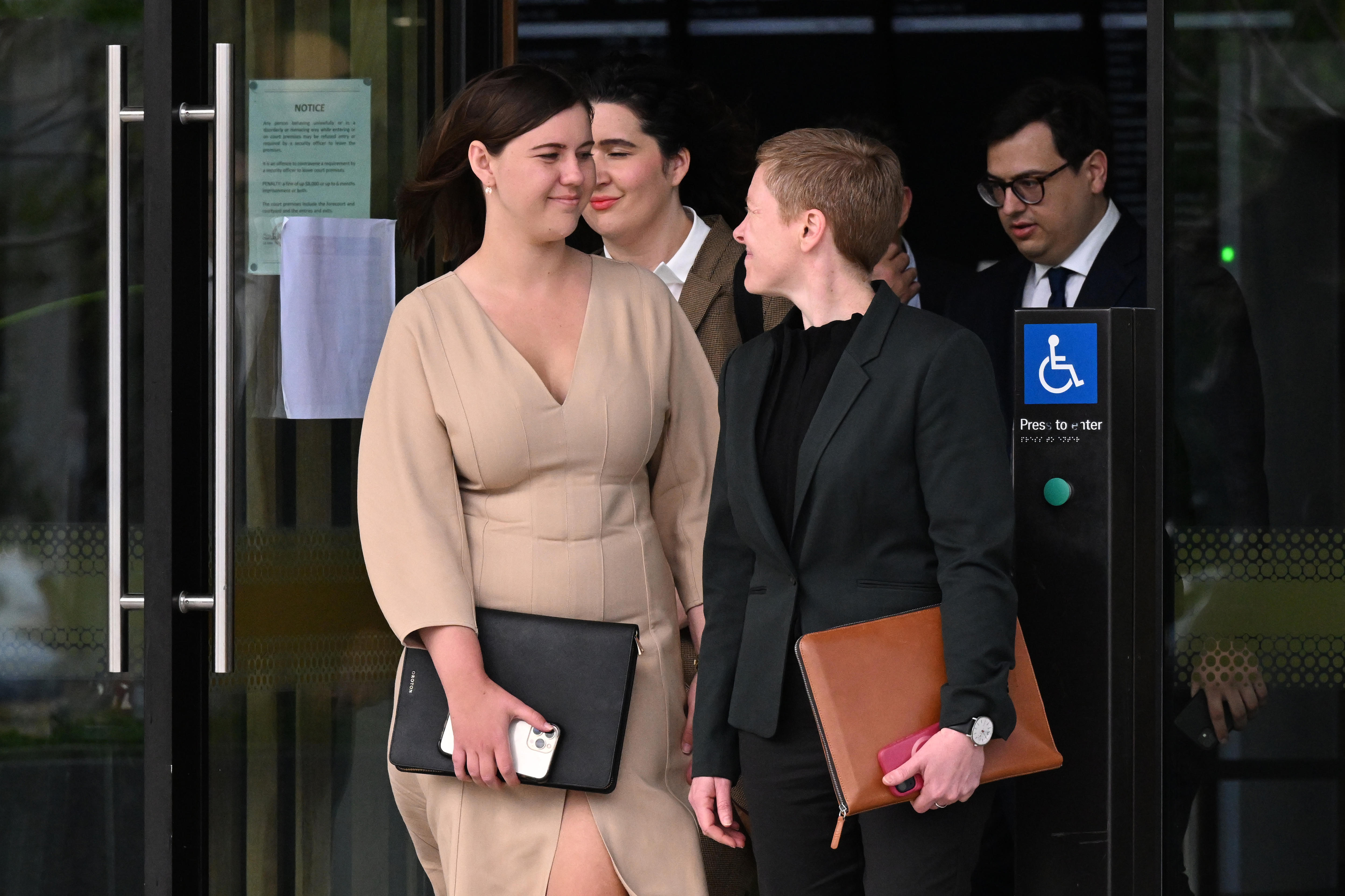 A woman in a beige dress walks out a door with a small group around her.