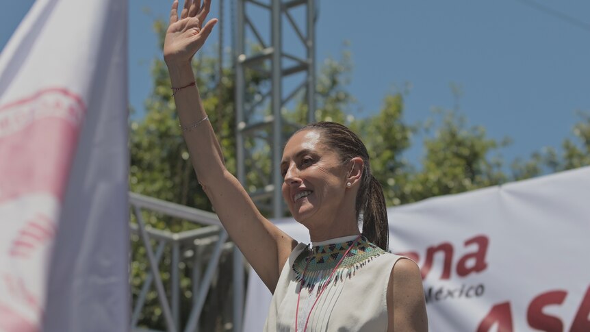 A female politician with brown hair waves to a crowd from a stage.