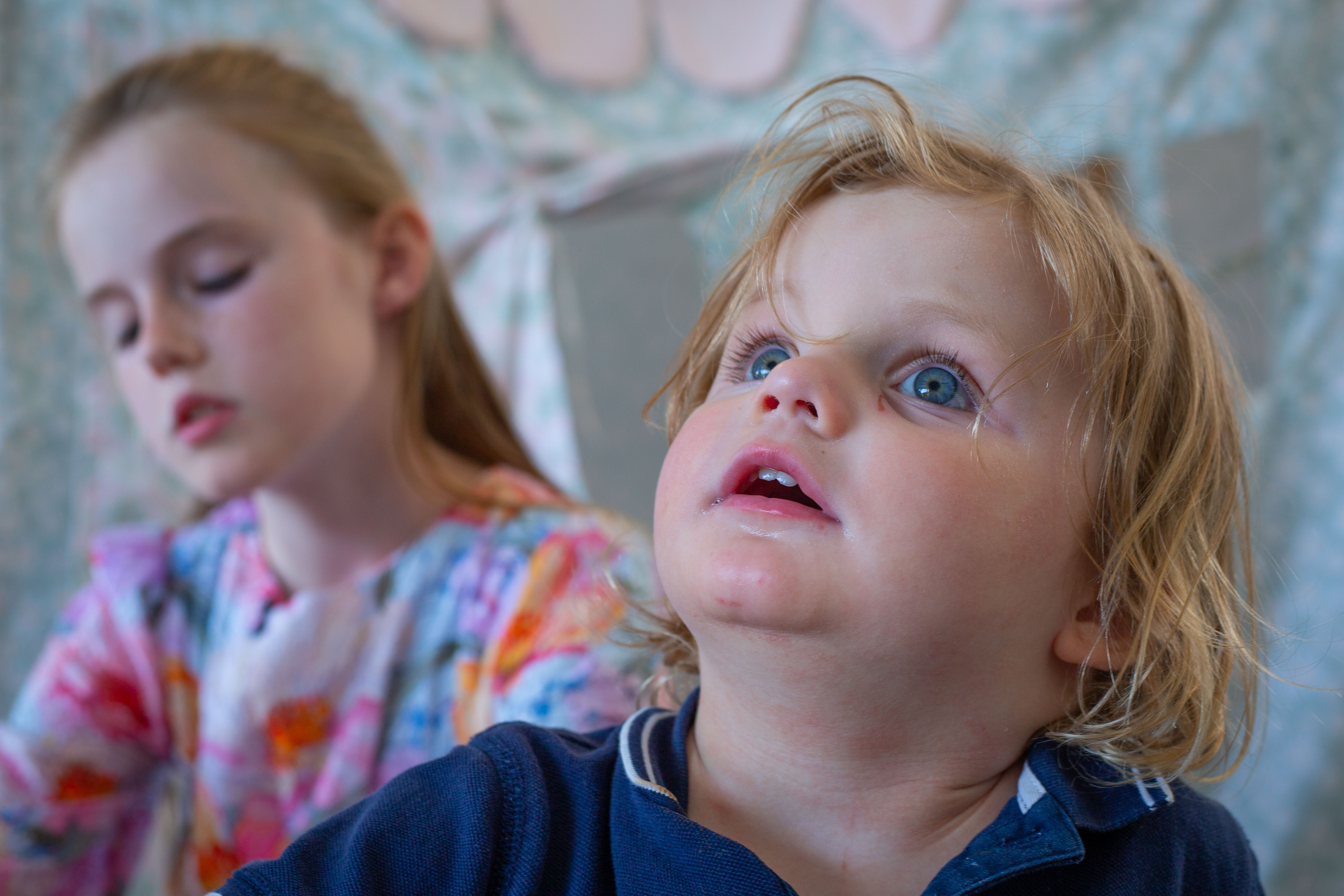 Un niño de tres años con el pelo rizado mira hacia arriba mientras su hermana mira al suelo al fondo.