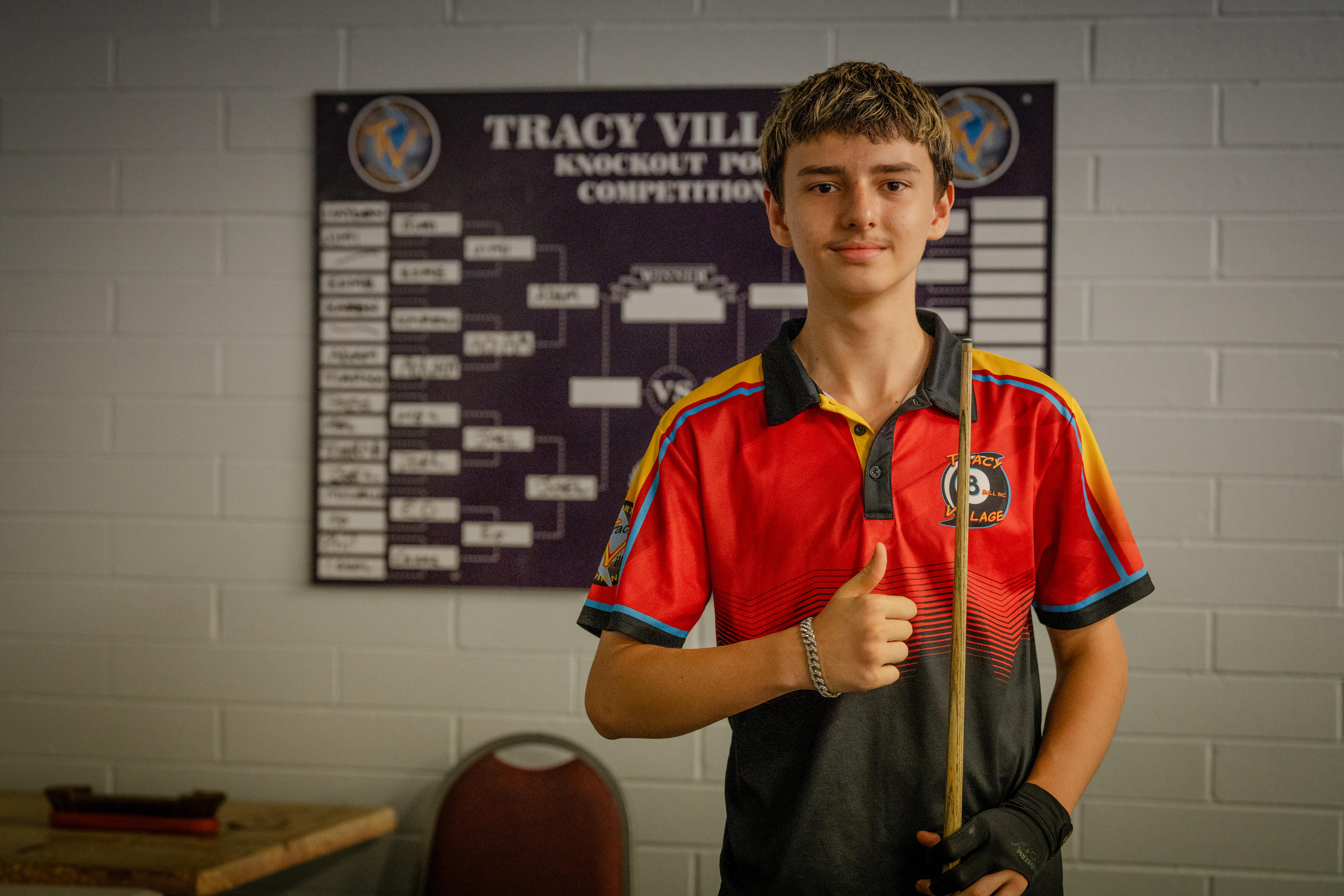 Adam Grace wears a local 8-ball club uniform, holds a pool cue and smiles at the camera while giving a thumbs up.