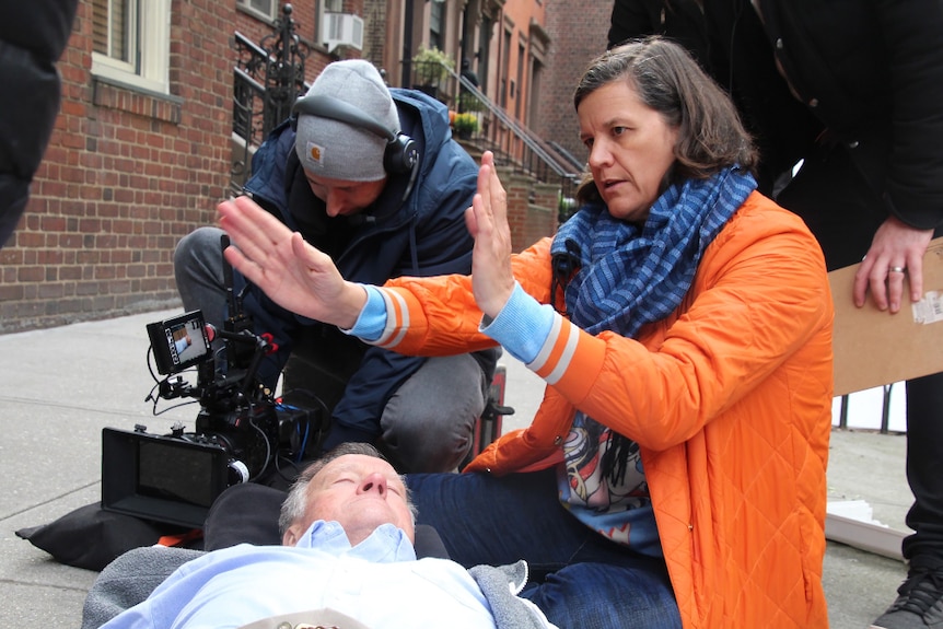 A camera operator kneels next to a brown haired woman in bright orange coat gesturing, both kneel over old man lying on ground.