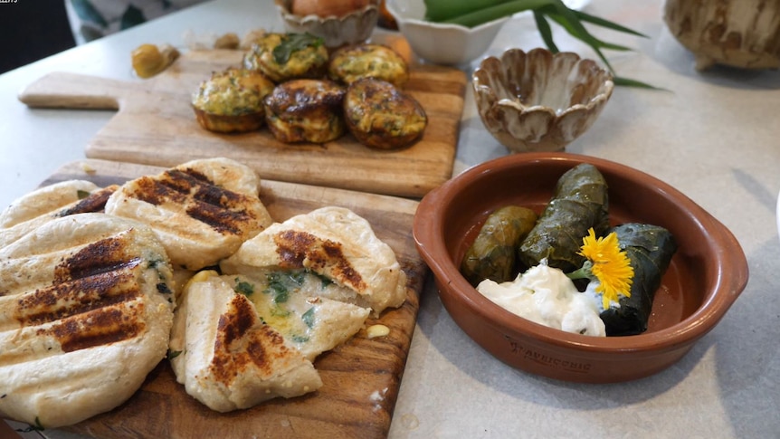 Bread with greens in the dough, mini quiche, dolmades laid out on boards and a brown clay dish.