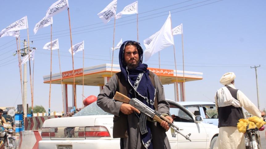 A man in robes holds a rifle outside a petrol station