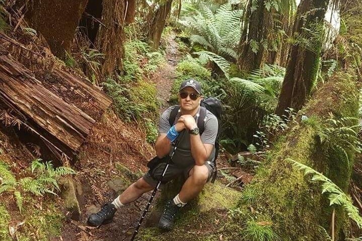 A male bushwalker rests on a rock on a track in a rainforest.