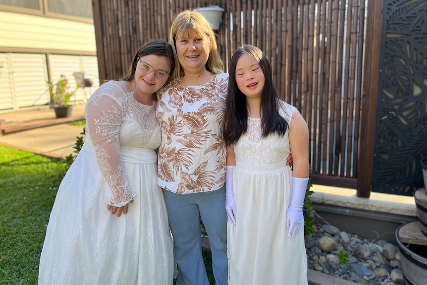 A woman stands between her two daughters in long white dresses.