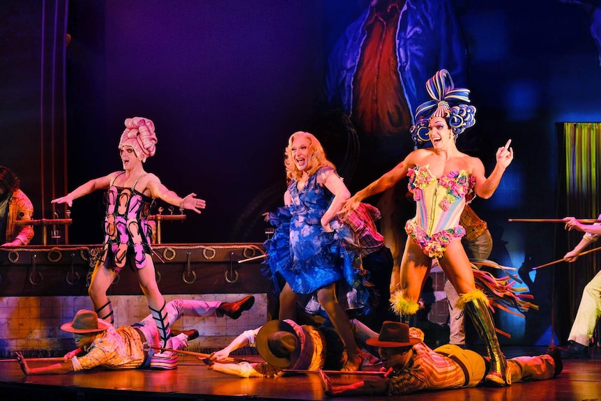 Two performers in colourful drag queen costumes and wigs flank Tony Sheldon, in dress and wig, in scene from the musical.