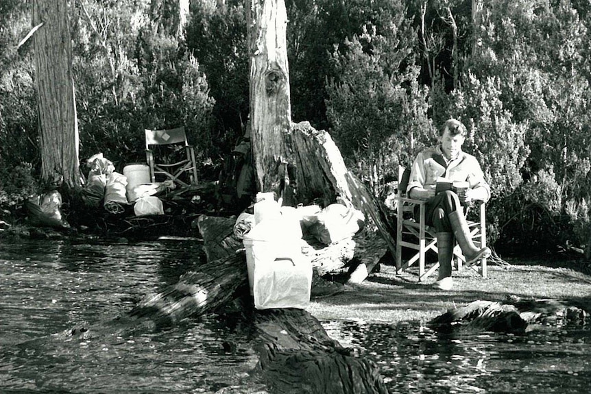 Black and white photo of a young man reading a book by a lake in the wilderness
