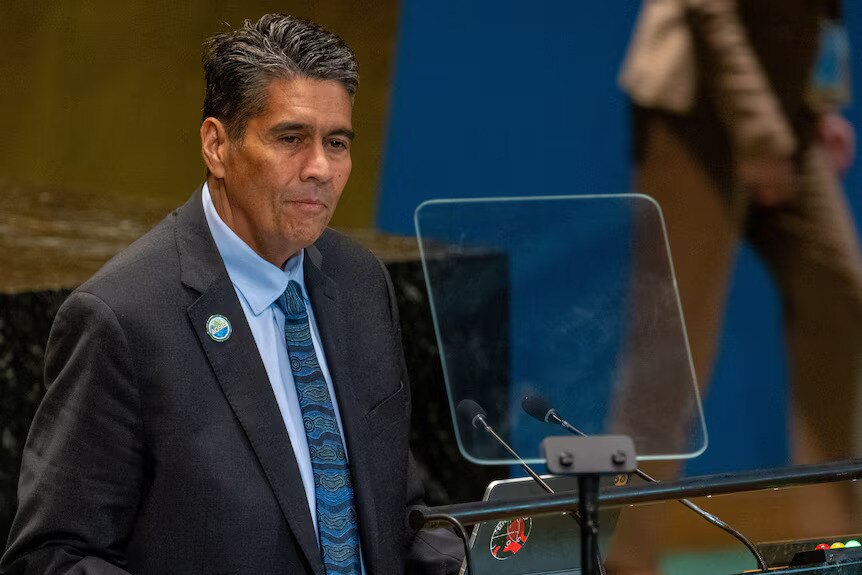 A man in a suit with a blue shirt and tie reads from a teleprompter 