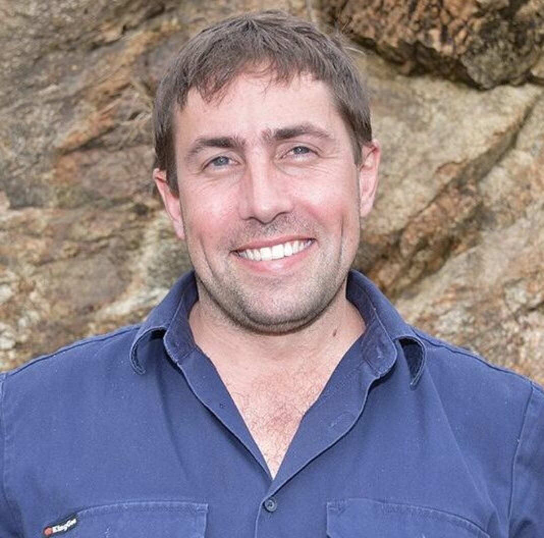 A young man with broad smile and blue shirt looks at camera.