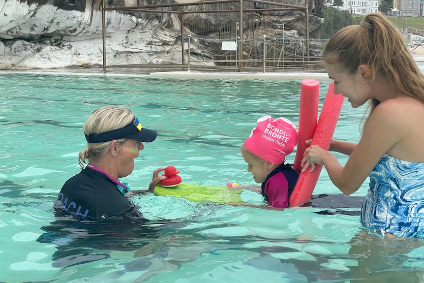 A woman in a cap smiles while a toddler swims holding a kickboard while wrapped in a pool noodle held by an adult woman