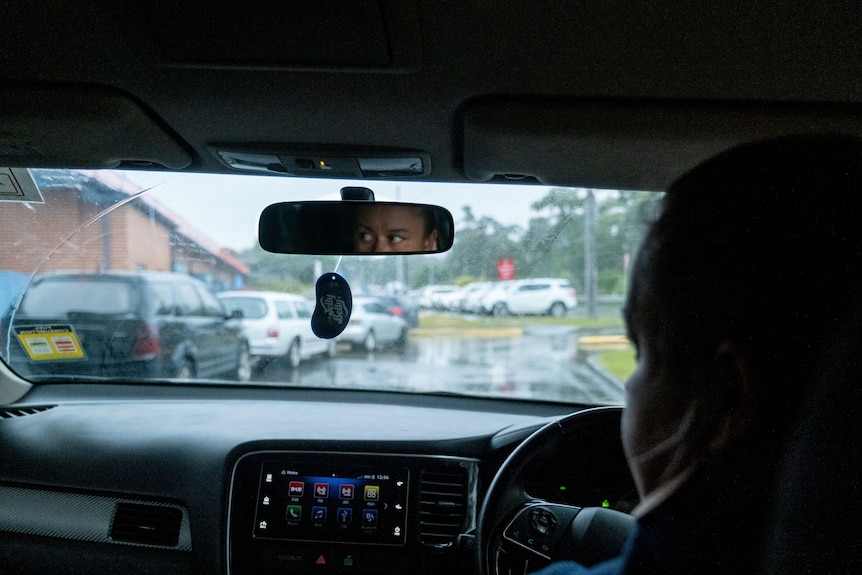 A woman drives her car into the parking lot of a hospital. Her determined eyes can be seen in the rear view mirror. 