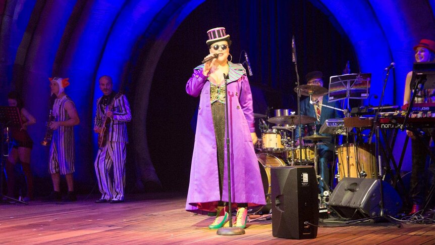 A woman in a coat and top hat sings with a 5 piece band