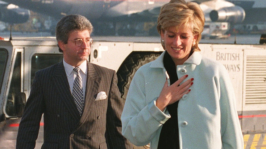 A woman in a light blue coat walks ahead of a bespectacled man in a suit with an attache case on the tarmac