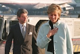 A woman in a light blue coat walks ahead of a bespectacled man in a suit with an attache case on the tarmac