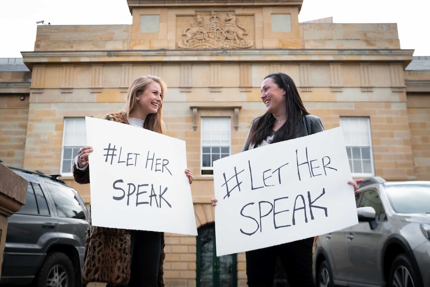 Two women with signs saying #LetHerSpeak, smiling at each other, Grace is on the left, Nina on the right