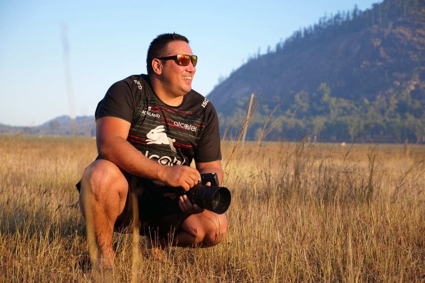 Trent White squats in the brown grass, wearing sunglasses, smiling, holding a camera, mountain behind.