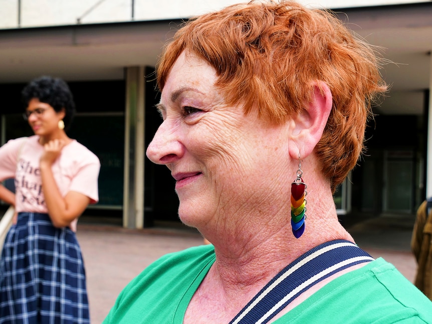 A woman smiles looking away from the camera, she wears rainbow earrings. 