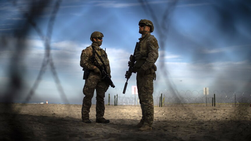 Two men wearing military-style uniforms and holding rifles stand in a desert environment, as seen through a roll of barbed wire.