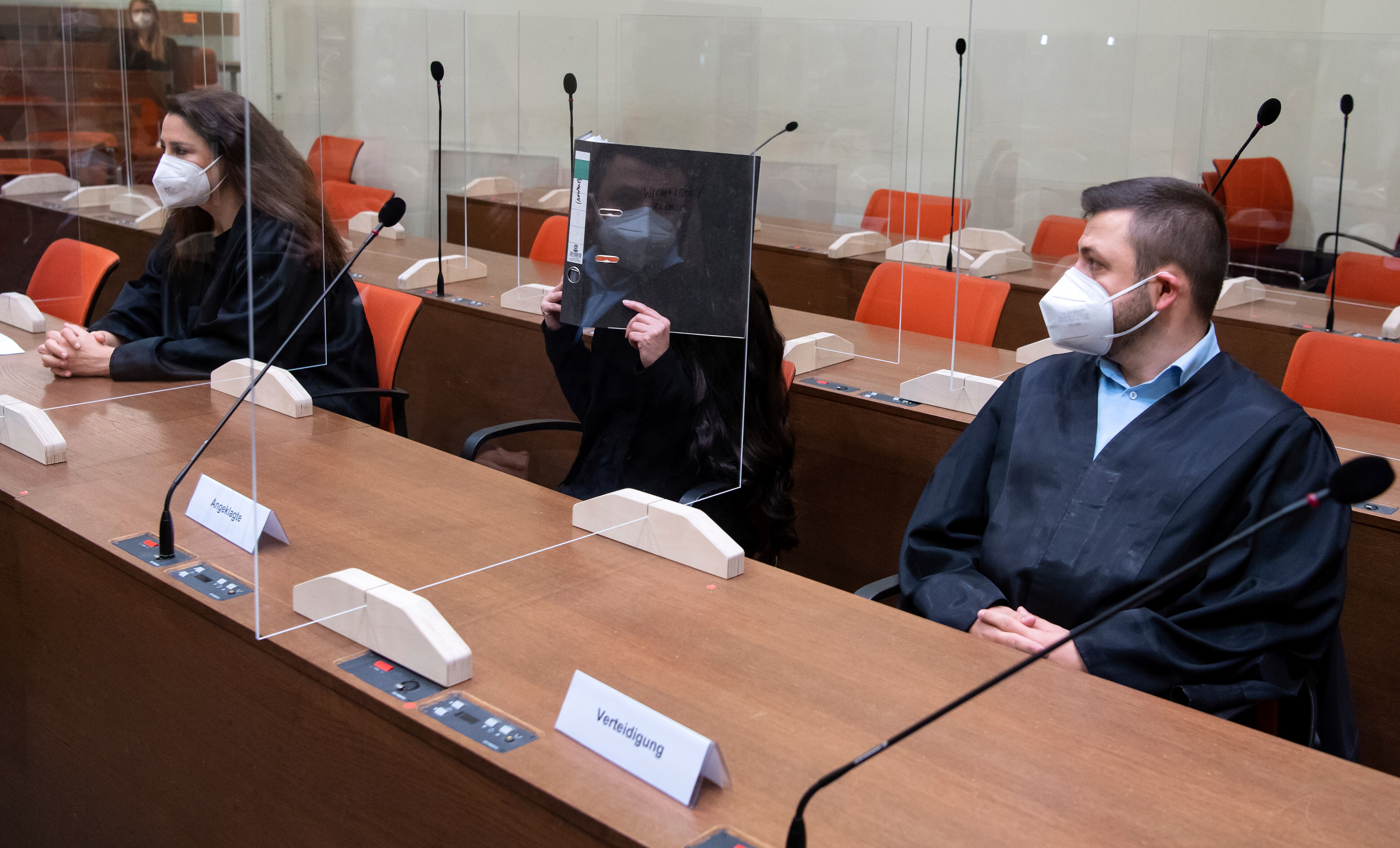 Sitting woman in black holds folder hiding face in court, while man next to her in mask looks on