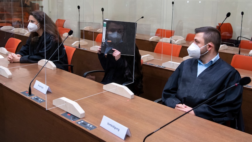 Sitting woman in black holds folder hiding face in court, while man next to her in mask looks on