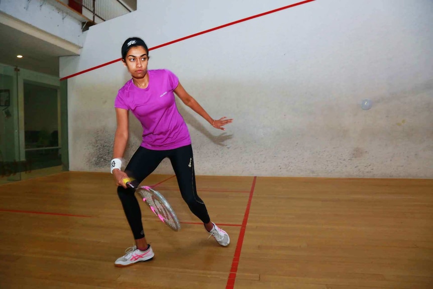 Woman in workout clothes hits a squash ball with her racquet on a squash court.