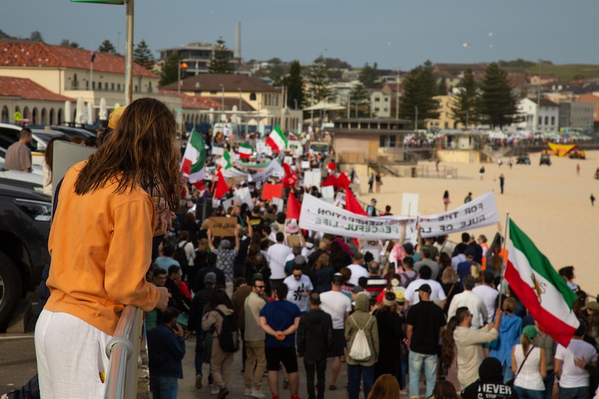 A woman in a yellow jumper looks on as a protest against Iran marches alongside Bondi Beach