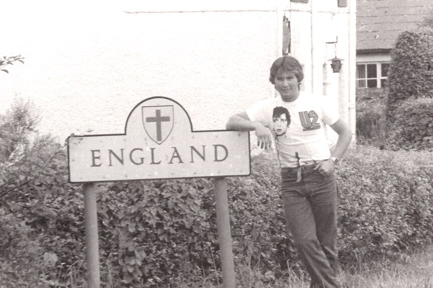 A young man leans against a sign that reads 'England'