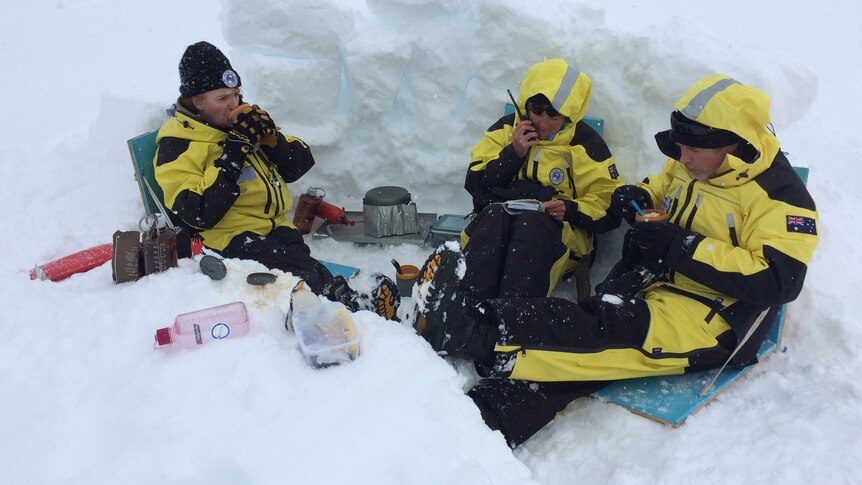 Three people in yellow and black snow gear in an icy dugout.