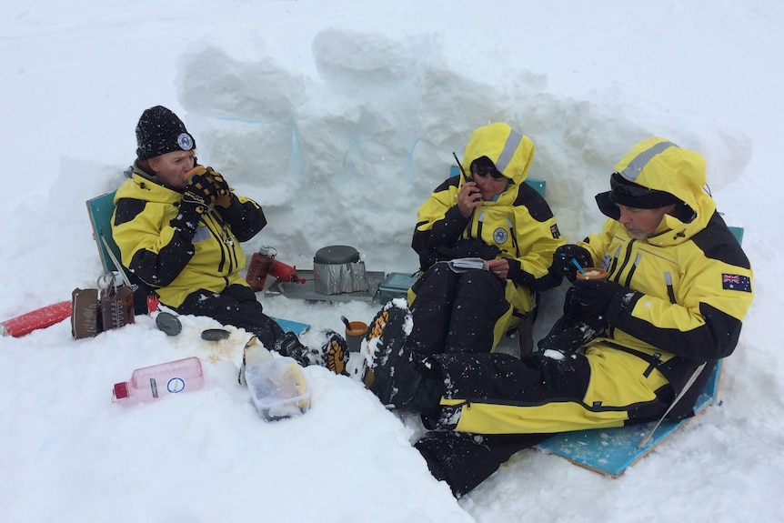 Three people in yellow and black snow gear in an icy dugout.