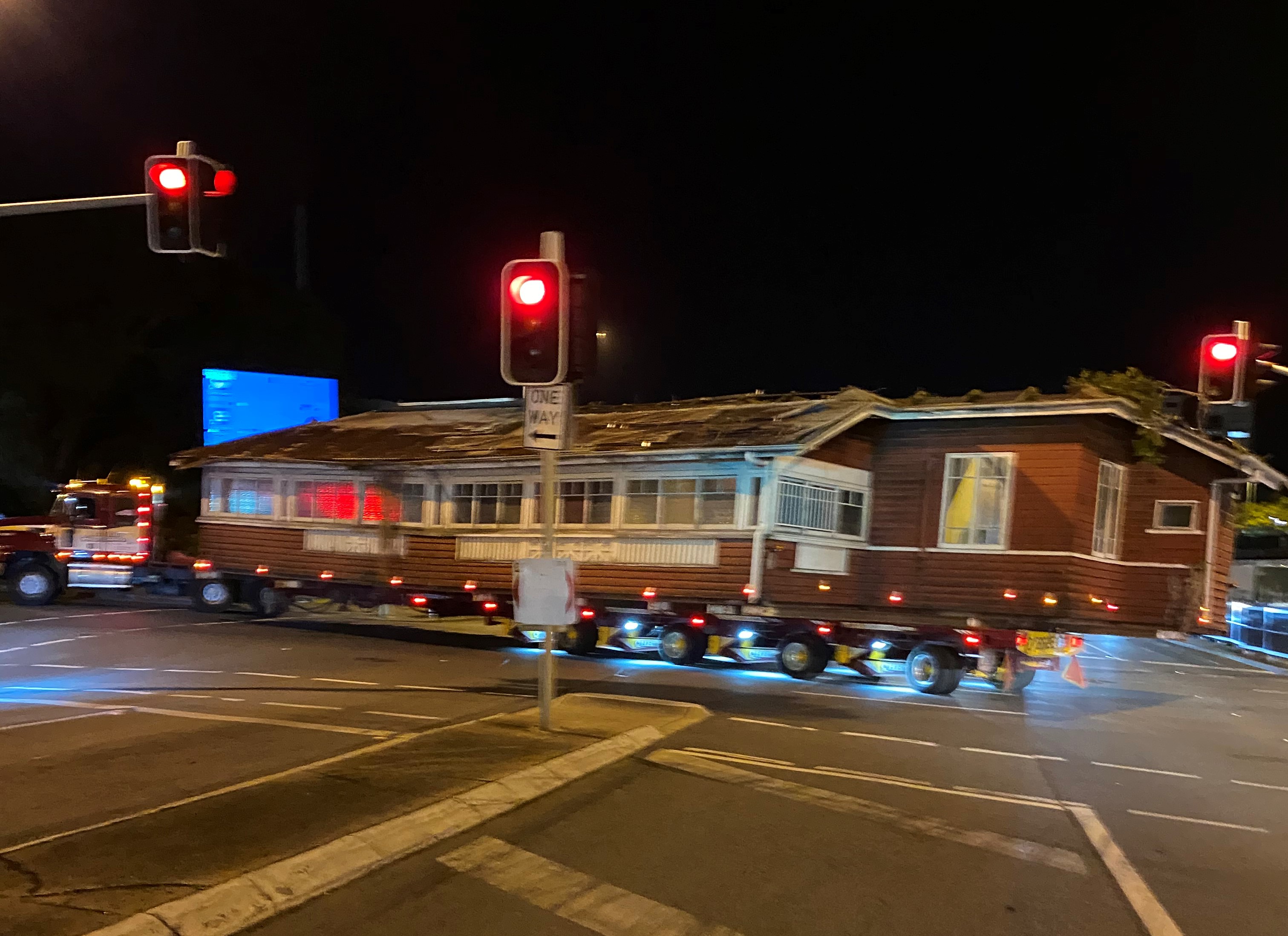 A red queenslander house on the back of a truck going through an intersection at night.