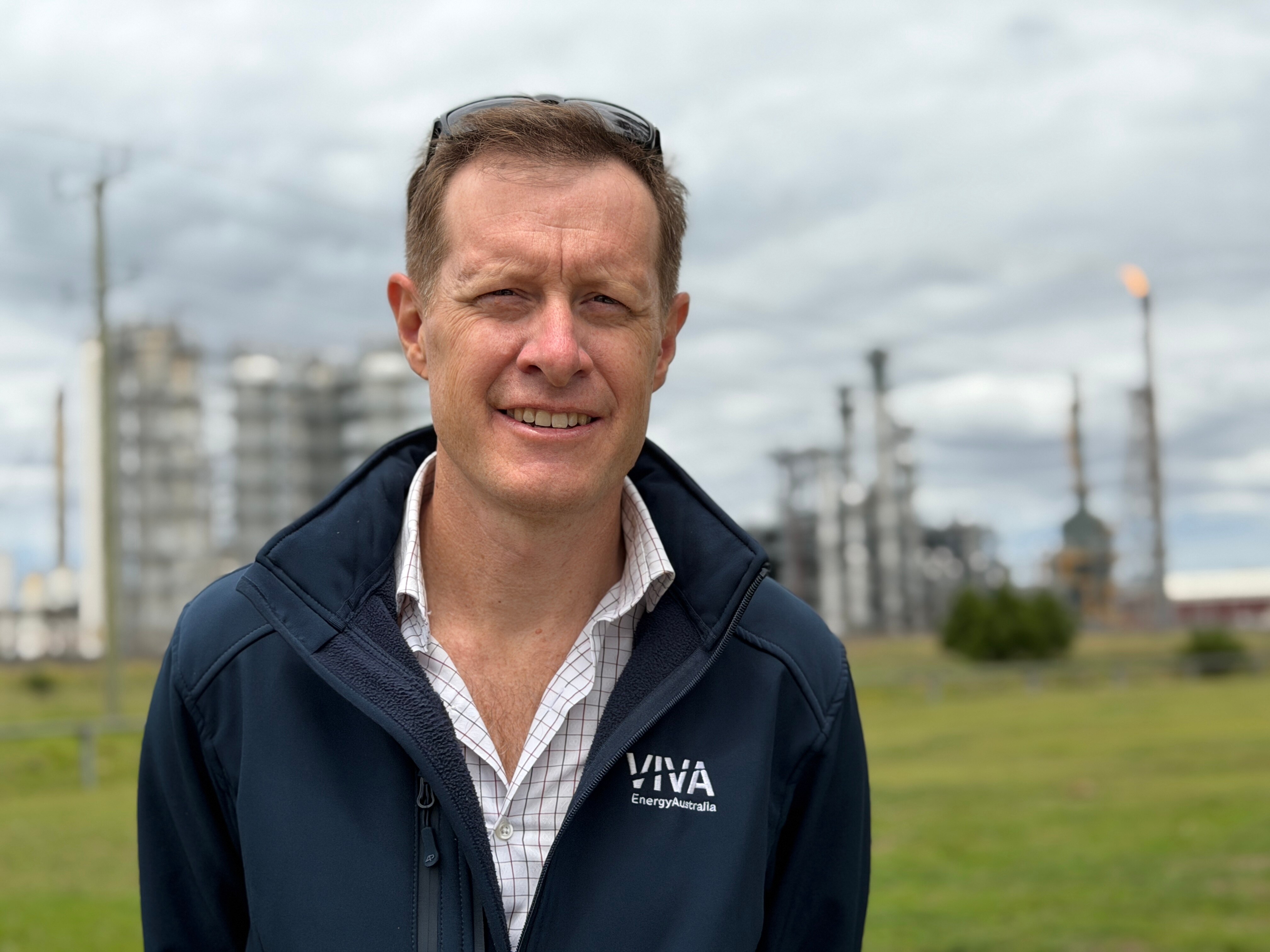 Un hombre con una chaqueta polar azul oscuro con una refinería de petróleo al fondo.
