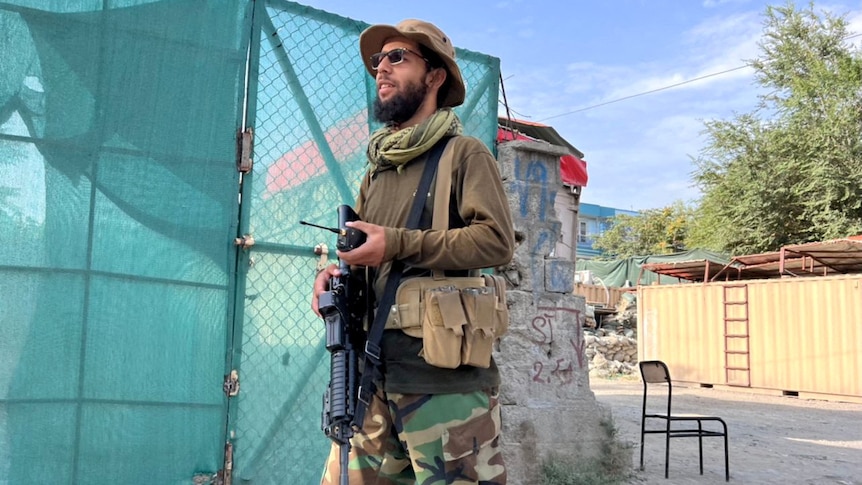 A man with a long beard, wearing camo gear and holding a giant long gun, stands next to an area covered in green tarps 