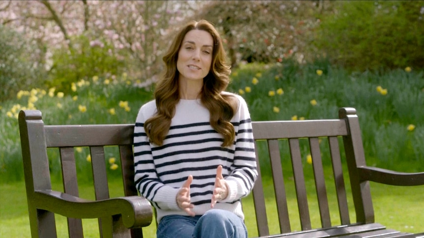 Princess Kate, in a striped jumper and jeans, sits on a wooden park bench in a garden addressing the camera. 