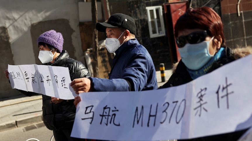 Two women and one man, in warm clothing, outside during the day, holding protest signs