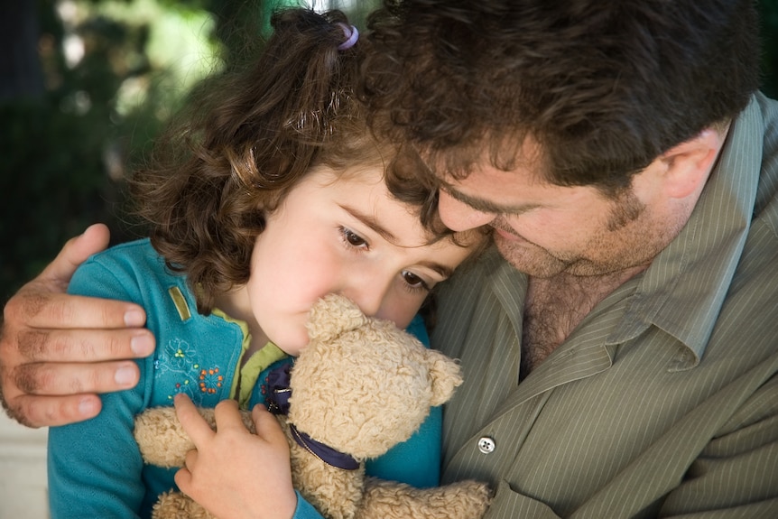 A man comforts a young girl holding a teddy bear