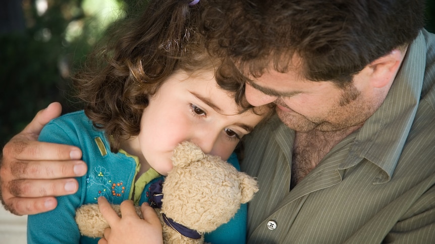 A man comforts a young girl holding a teddy bear