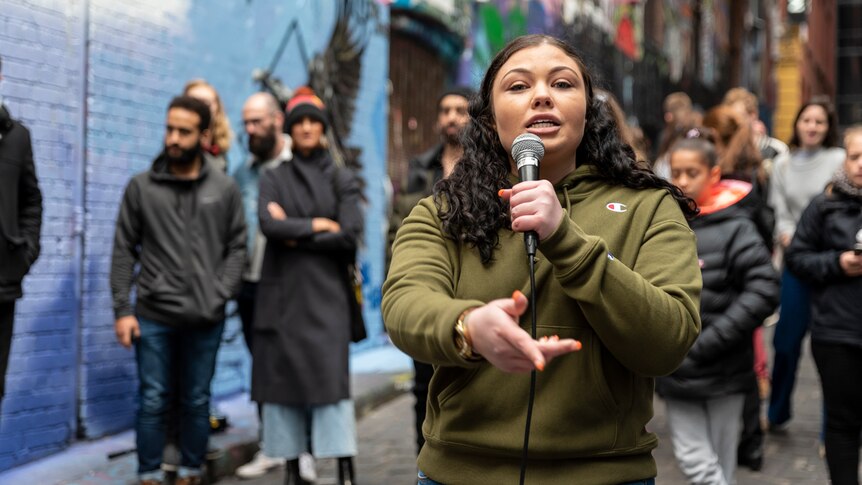 Young woman in olive green hoodie with long brown hair holds mic and sings looking at camera, outdoor in laneway with passersby.