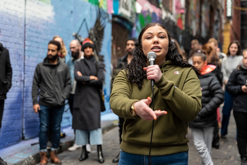 Young woman in olive green hoodie with long brown hair holds mic and sings looking at camera, outdoor in laneway with passersby.