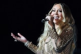 Pop star Mariah Carey sings into a microphone with a smile, wearing a glittering gown.