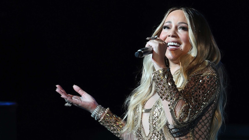 Pop star Mariah Carey sings into a microphone with a smile, wearing a glittering gown.